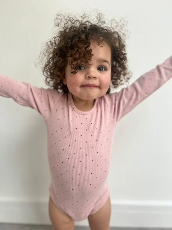 A young girl with curly hair and blue eyes standing in front of a white wall, wearing a pink long-sleeved onesie with small polka dots, arms raised up. Modelling for leading child model and talent agency SocityUK based in London and Manchester.
