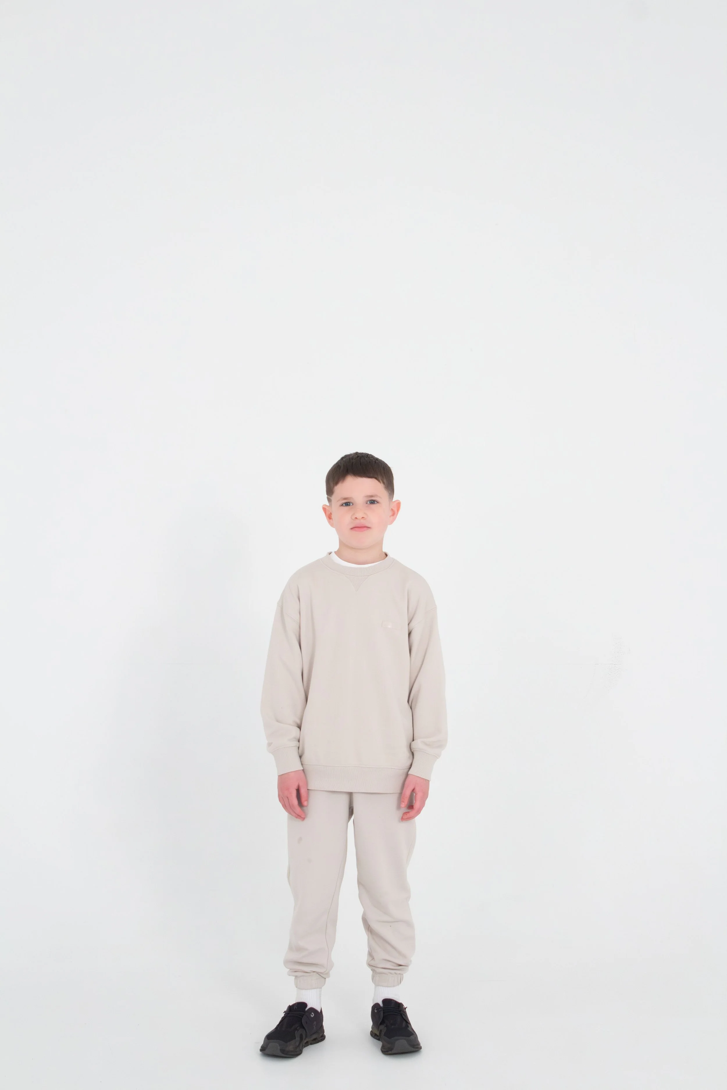 A young boy stands with a neutral expression in a plain white room, wearing a beige sweatshirt, matching beige sweatpants, and black sneakers.