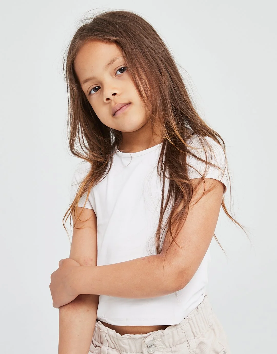 A young girl with long brown hair, wearing a white t-shirt and beige pants, standing with arms crossed against a plain background.
