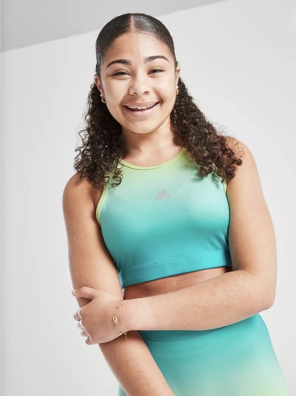 A young woman with curly dark hair in a ponytail, smiling, wearing a green and teal athletic crop top and matching skirt, standing against a plain light background.