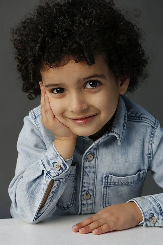 A young boy with curly hair smiling, resting his head on his hand, wearing a denim jacket, against a dark gray background.