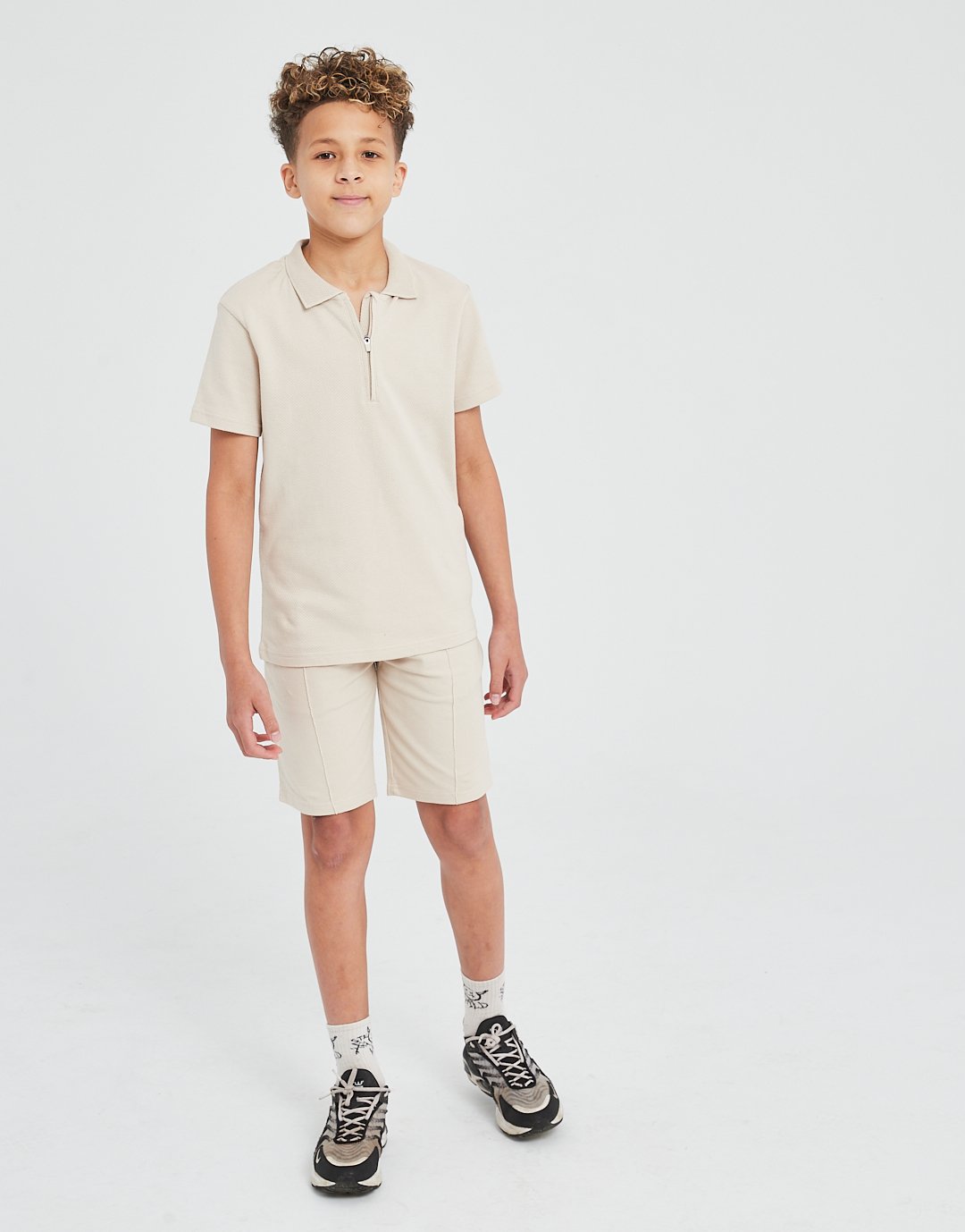 Young boy standing against a plain white background, wearing a beige polo shirt with a zipper, matching beige shorts, white socks with black design, and black sneakers.