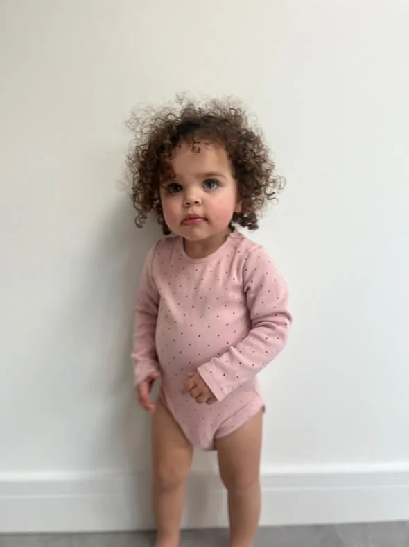 A young girl with curly hair wearing a pink long-sleeve shirt and matching shorts standing against a white wall. Modelling for leading child model and talent agency SocityUK based in London and Manchester.