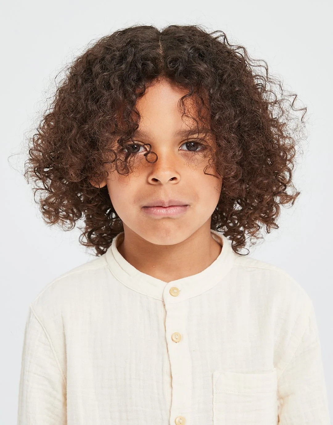 Close-up portrait of a young boy with curly brown hair, wearing a cream-colored button-up shirt, standing against a white background.