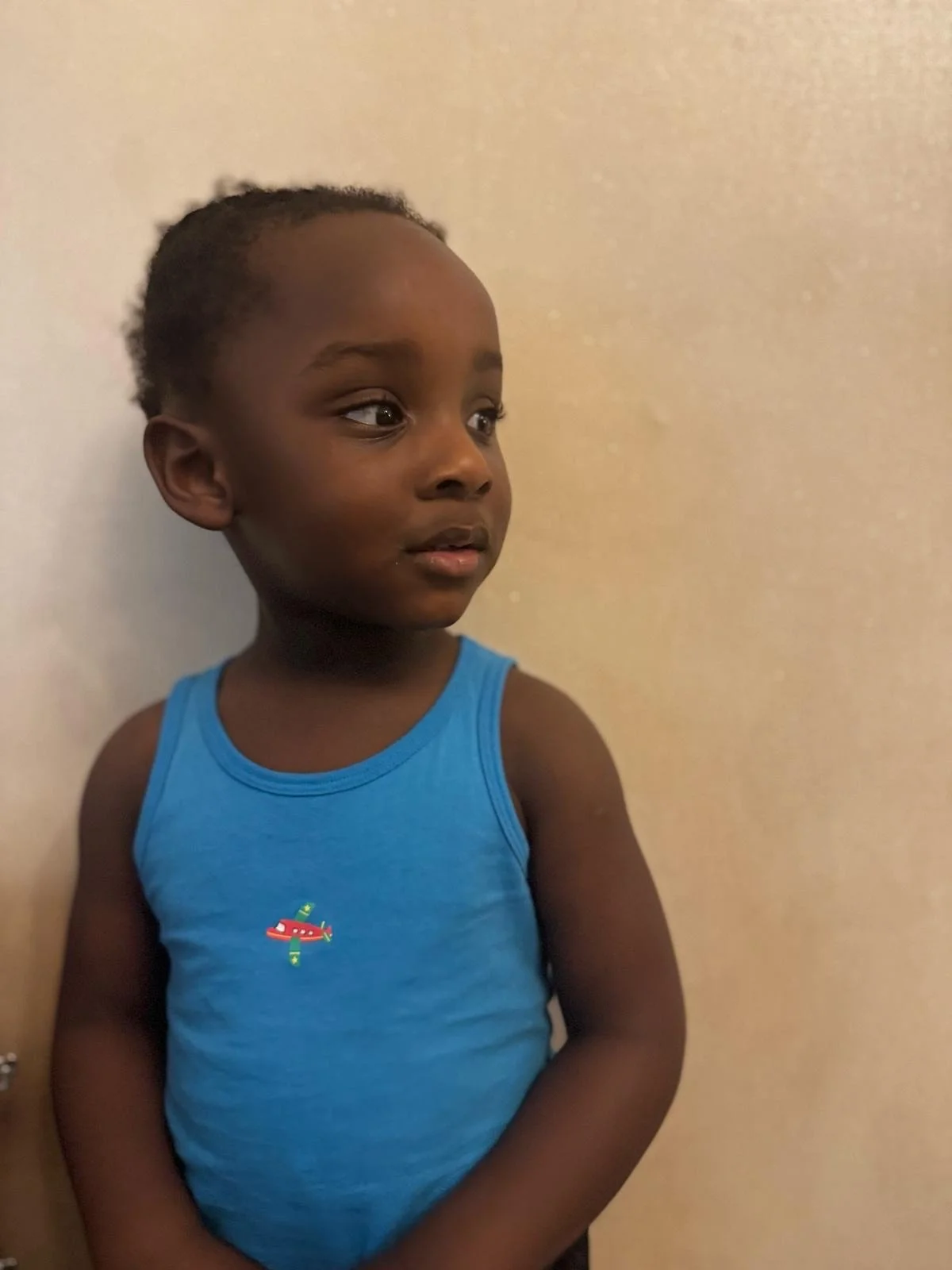 A young child with short curly hair wearing a blue tank top with a small red airplane design, standing against a plain beige wall. Modelling for leading child model and talent agency SocityUK based in London and Manchester.