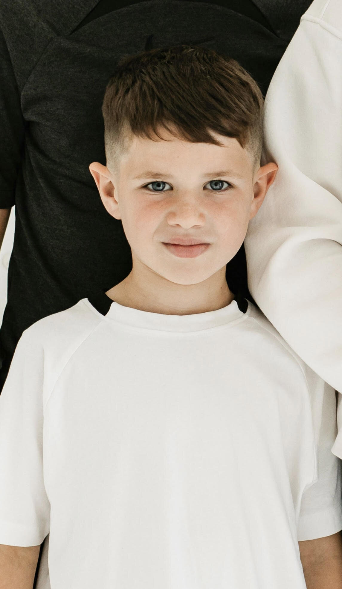 Close-up of a young boy with blue eyes, light skin, short brown hair, lying on a bed with a white and black shirt, looking at the camera.