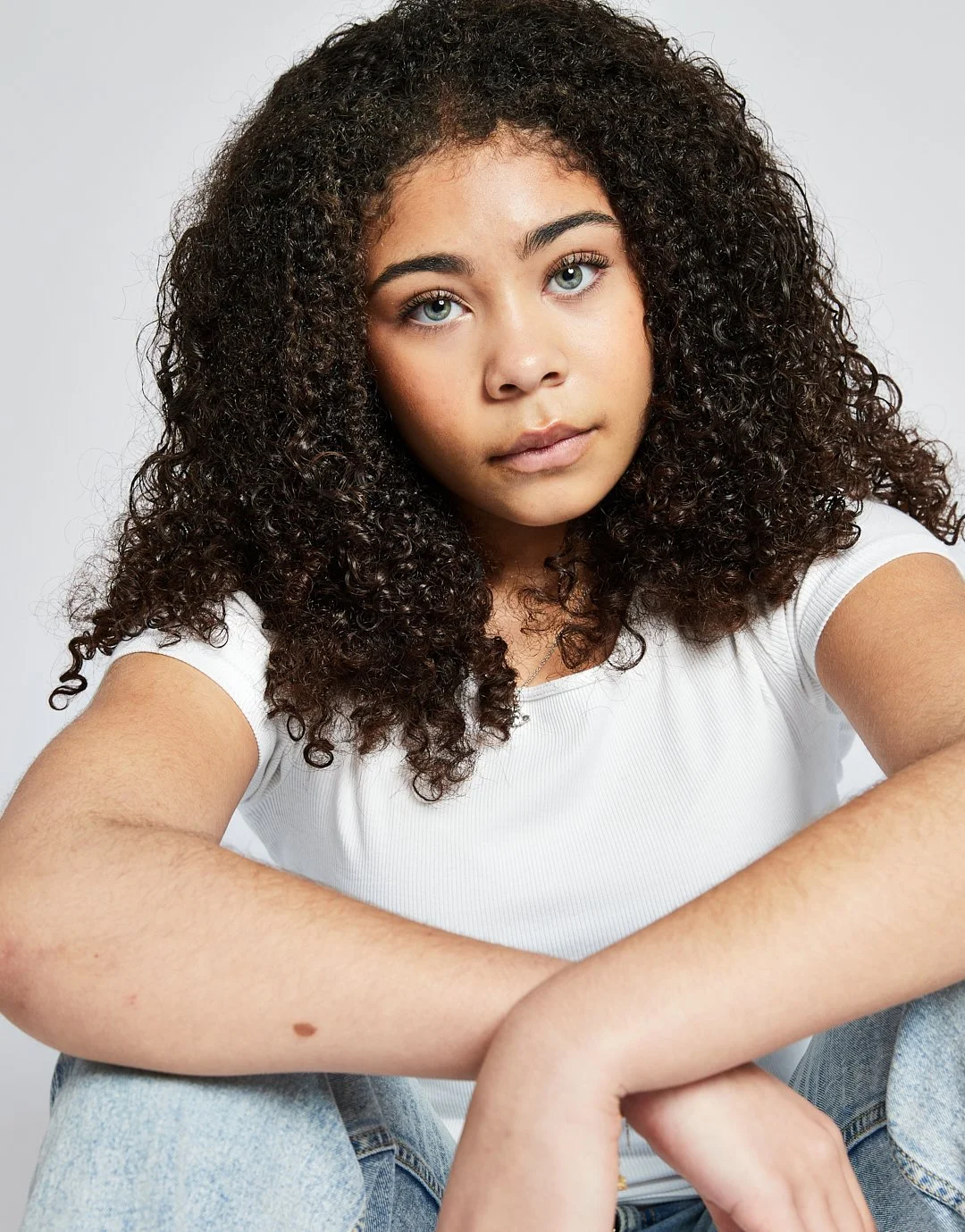 A young woman with curly dark hair and blue eyes wearing a white t-shirt, sitting with arms crossed, looking directly at the camera against a plain light background.
