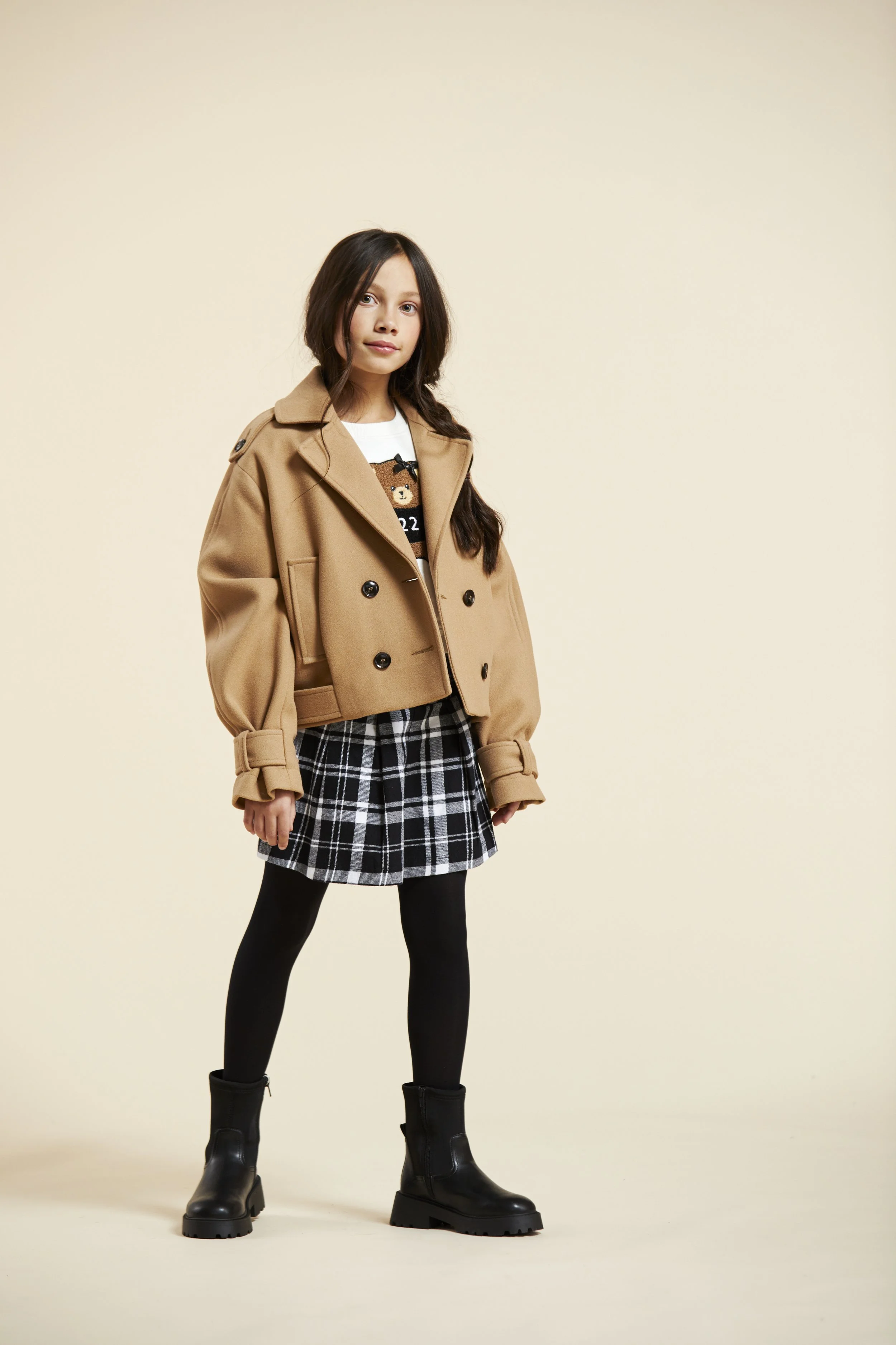 Young girl wearing a tan cropped coat, plaid skirt, black tights, and black boots standing against a plain beige background. Modelling for leading child model and talent agency SocityUK based in London and Manchester.