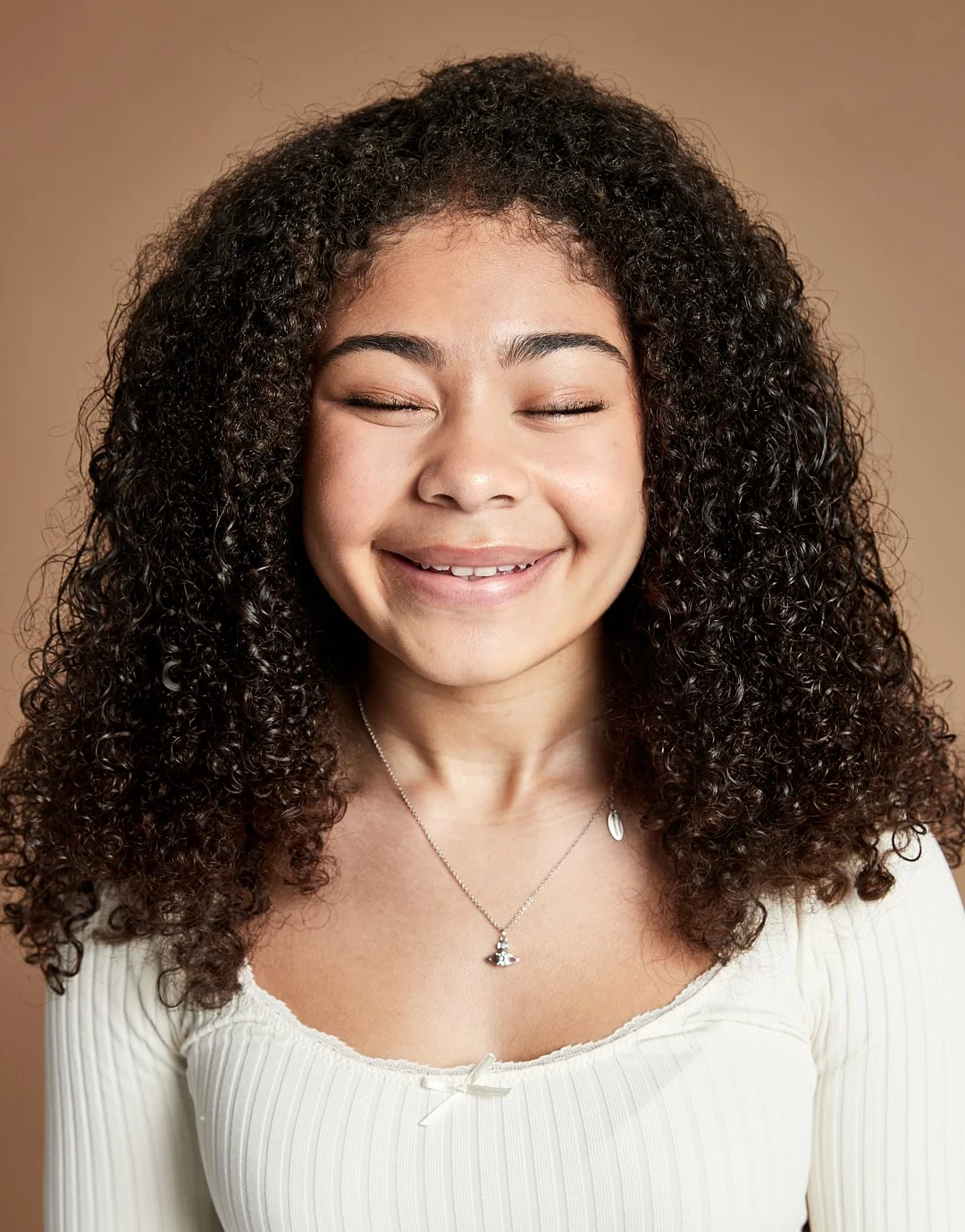 A young woman with curly hair smiling with eyes closed, wearing a white top and a silver necklace, against a neutral background.