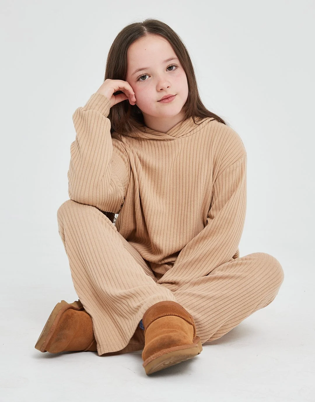 A young girl with brown hair, wearing a beige ribbed hoodie and matching pants, sits cross-legged on the floor with her left elbow resting on her knee and her head leaning on her hand. She has a neutral expression and is wearing tan boots.