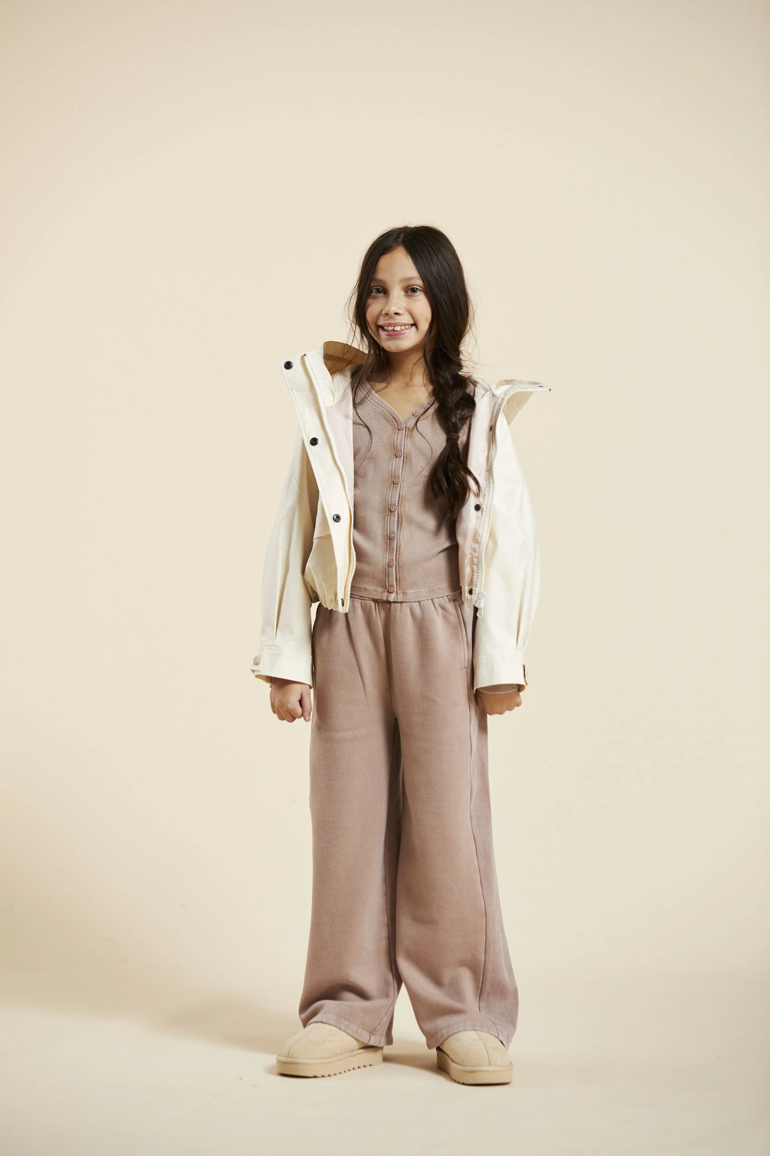 A young girl smiling, wearing a beige jumpsuit, a light-colored jacket, and beige shoes, standing against a plain light background. Modelling for leading child model and talent agency SocityUK based in London and Manchester.
