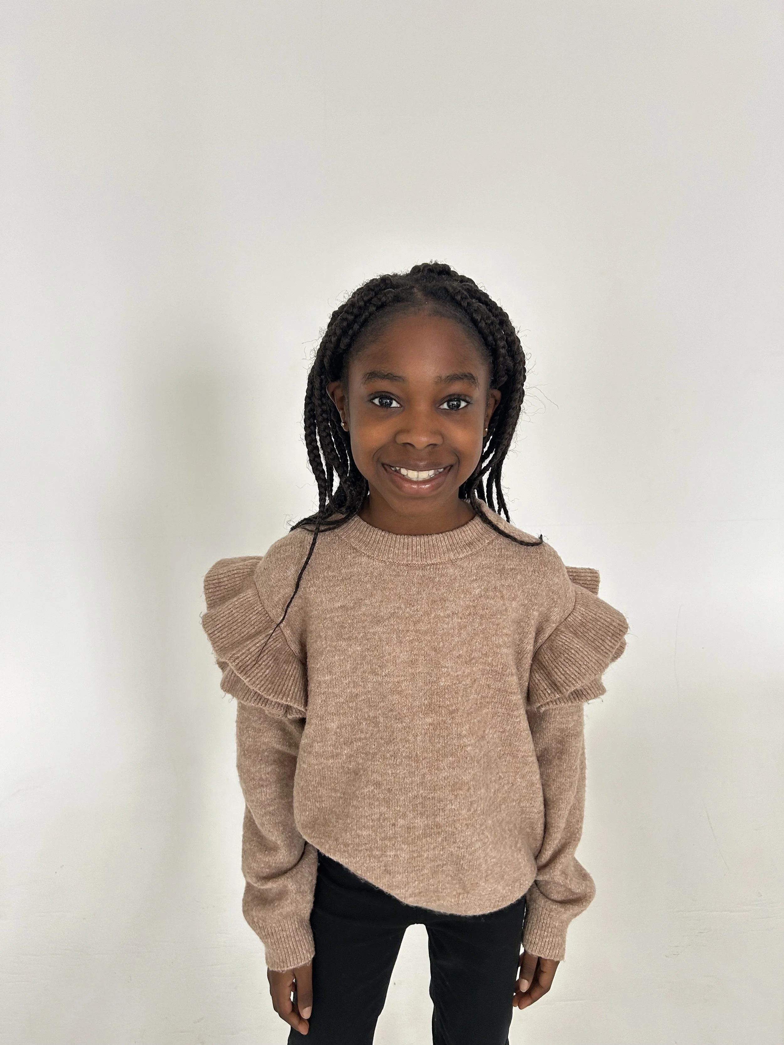 A young girl with braided hair smiling, wearing a beige sweater with ruffled shoulders and black pants, standing against a plain white background.