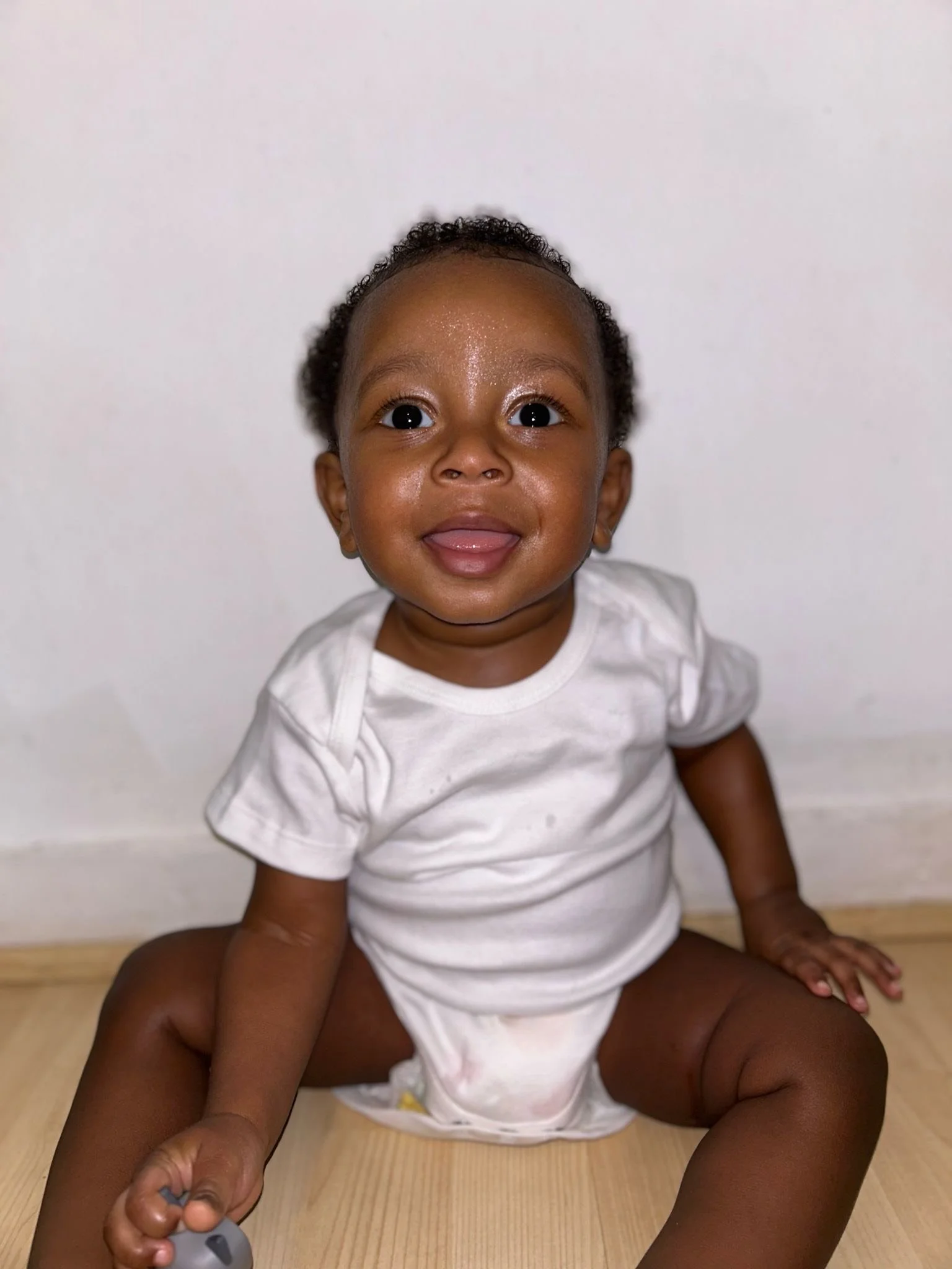 A happy baby with dark skin and curly hair sitting on a wooden floor against a white wall, smiling and looking at the camera. Modelling for leading child model and talent agency SocityUK based in London and Manchester.