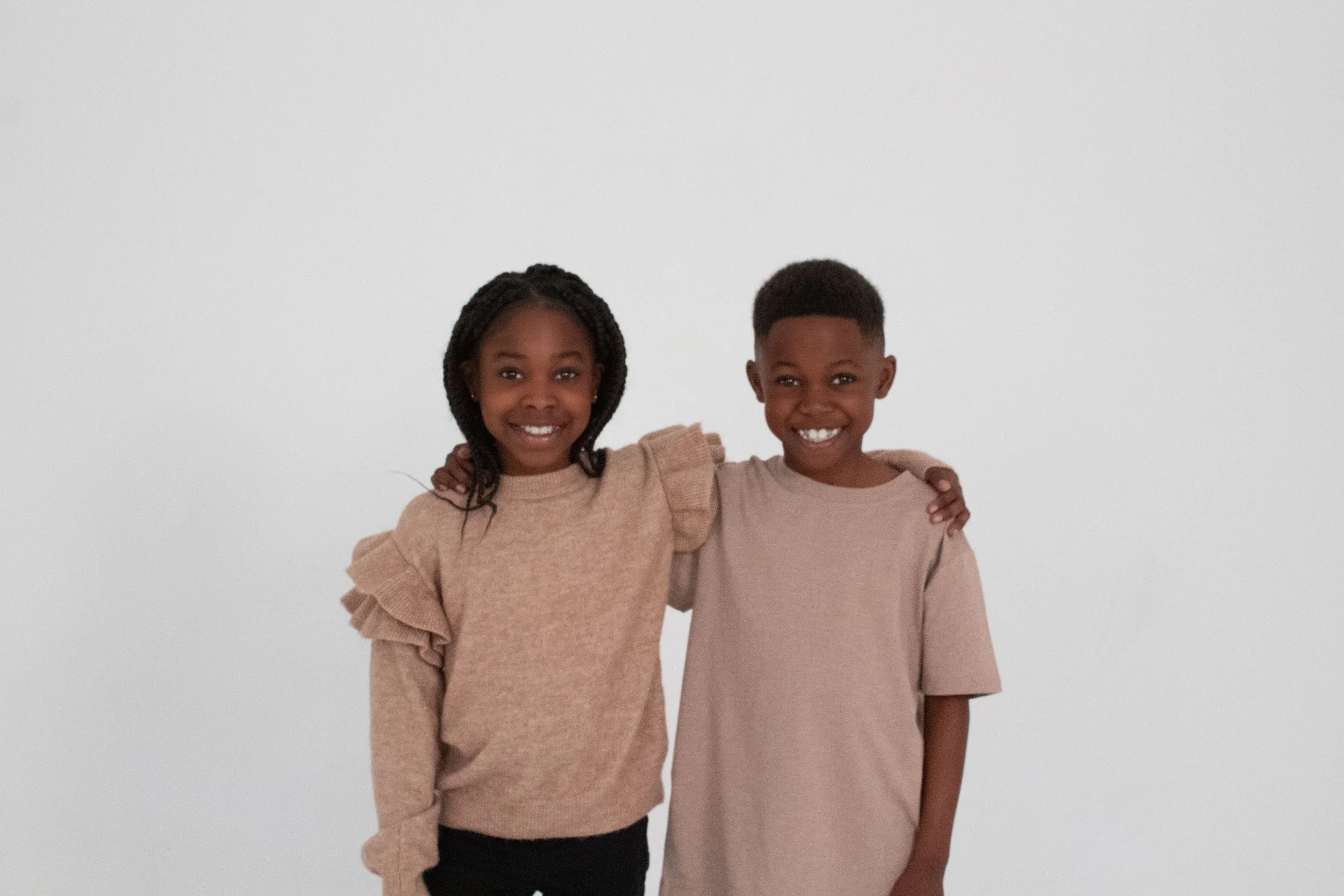 A girl and a boy standing close together, smiling with their arms around each other's shoulders, against a plain white background.