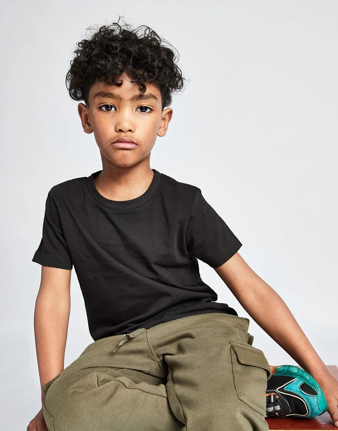 A young boy with curly black hair, wearing a black t-shirt and khaki cargo pants, sitting against a plain light gray background.Modelling for leading child model and talent agency SocityUK based in London and Manchester.