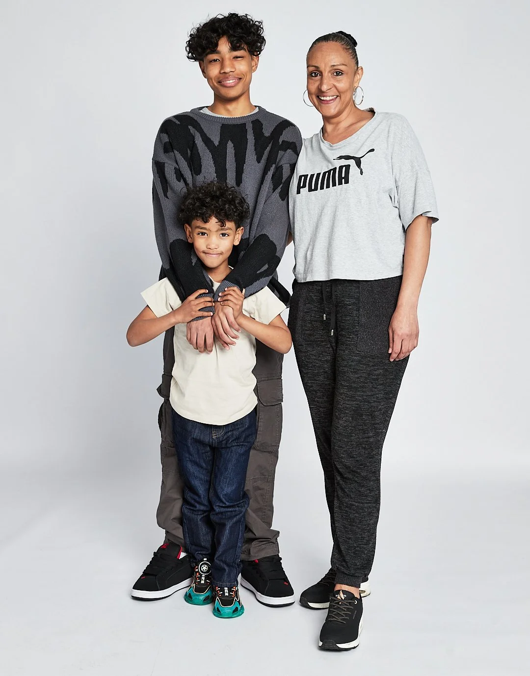 A woman and two boys standing together, smiling, against a plain light background. The woman is wearing a gray Puma t-shirt and black pants, the older boy is wearing a gray and black patterned sweater and gray pants, and the younger boy is in a cream