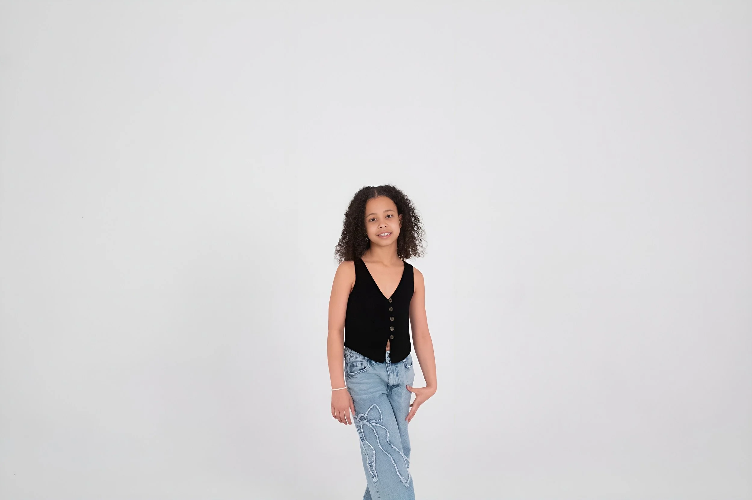 A young girl with curly hair wearing a black sleeveless top and light jeans standing against a plain white background.
