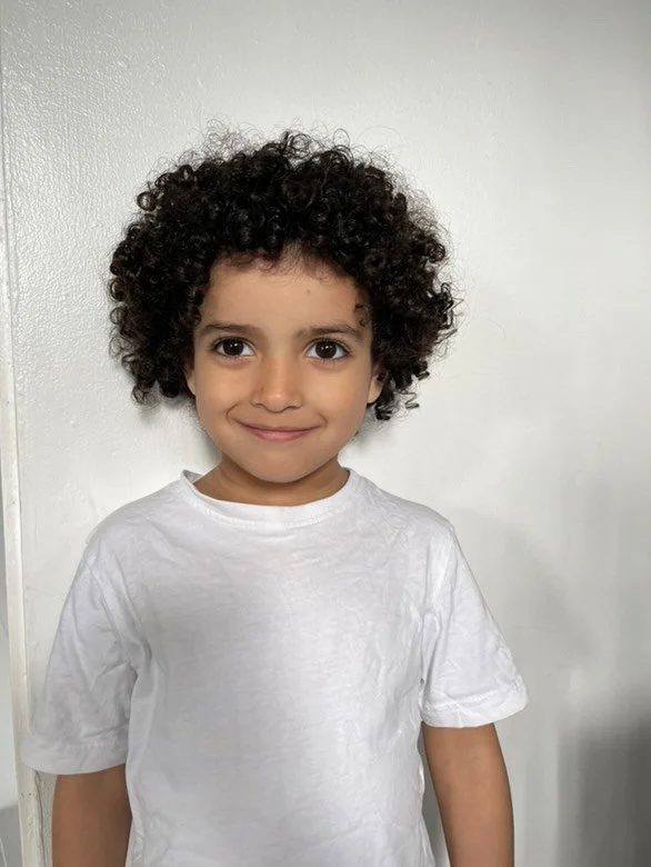 A young child with curly dark hair wearing a white t-shirt, smiling at the camera against a plain white wall.