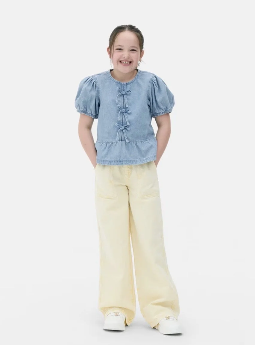 Young girl smiling, wearing a blue top with puffed sleeves and bows, yellow pants, and white shoes, standing against a white background.