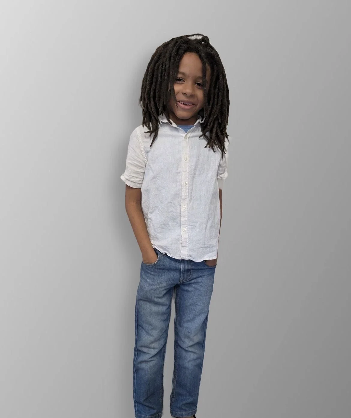 A young boy with long dreadlocks standing against a gray background, smiling, wearing a white button-up shirt and blue jeans.