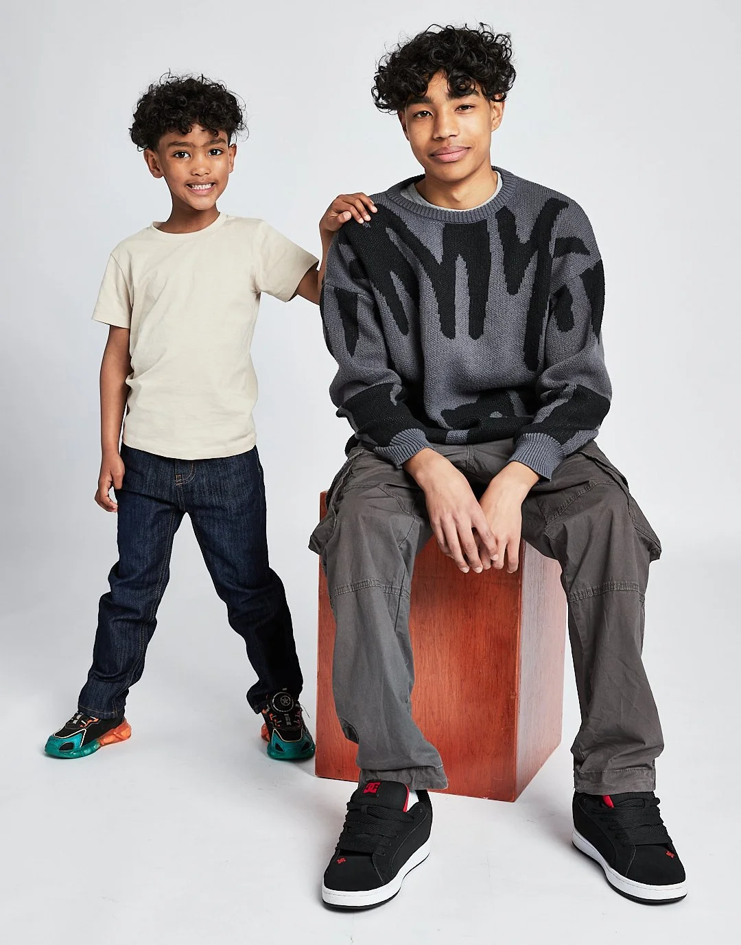A young boy with curly hair and a pastel T-shirt standing next to an older teenage boy with curly hair seated on a wooden block, both smiling in front of a plain white background. Modelling for leading child model and talent agency SocityUK based in 