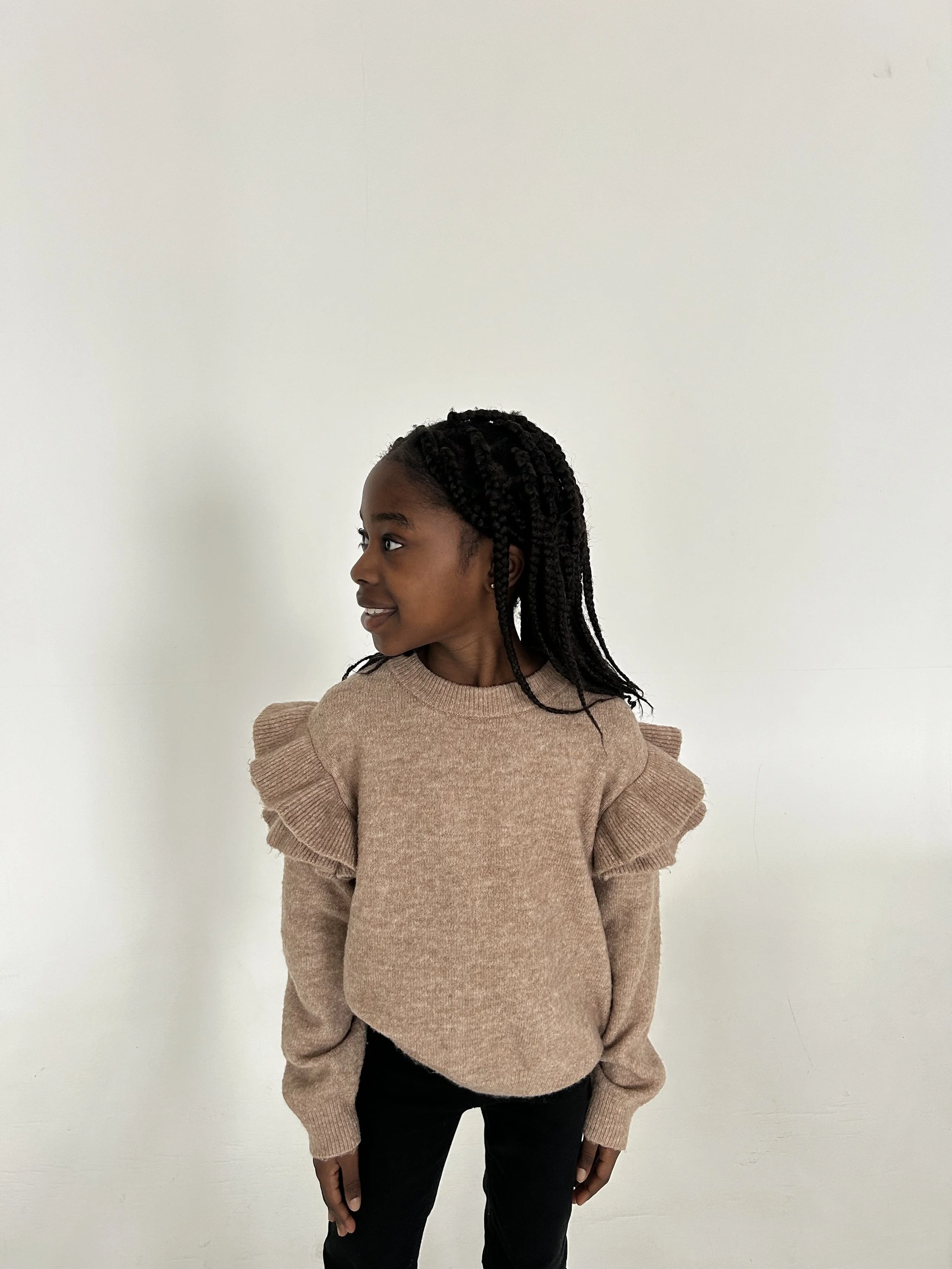 Young girl with braided hair wearing a beige sweater with ruffled shoulders and black pants, standing against a plain white wall, looking to her left. Model for leading Manchester and London based child model agency Socityuk.