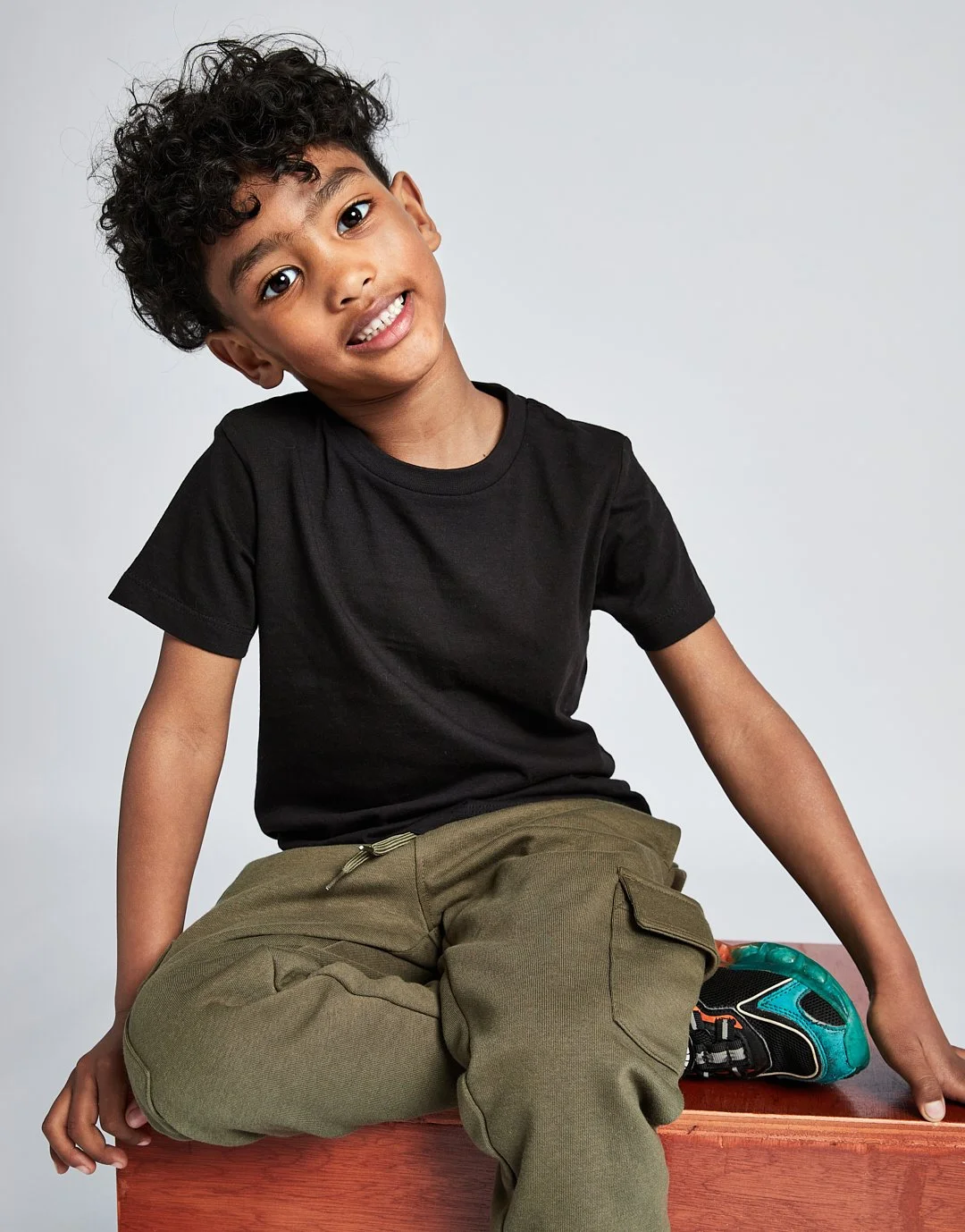 A young boy with curly hair wearing a black t-shirt and khaki pants sitting on a wooden surface against a plain background, looking at the camera with a slight head tilt.