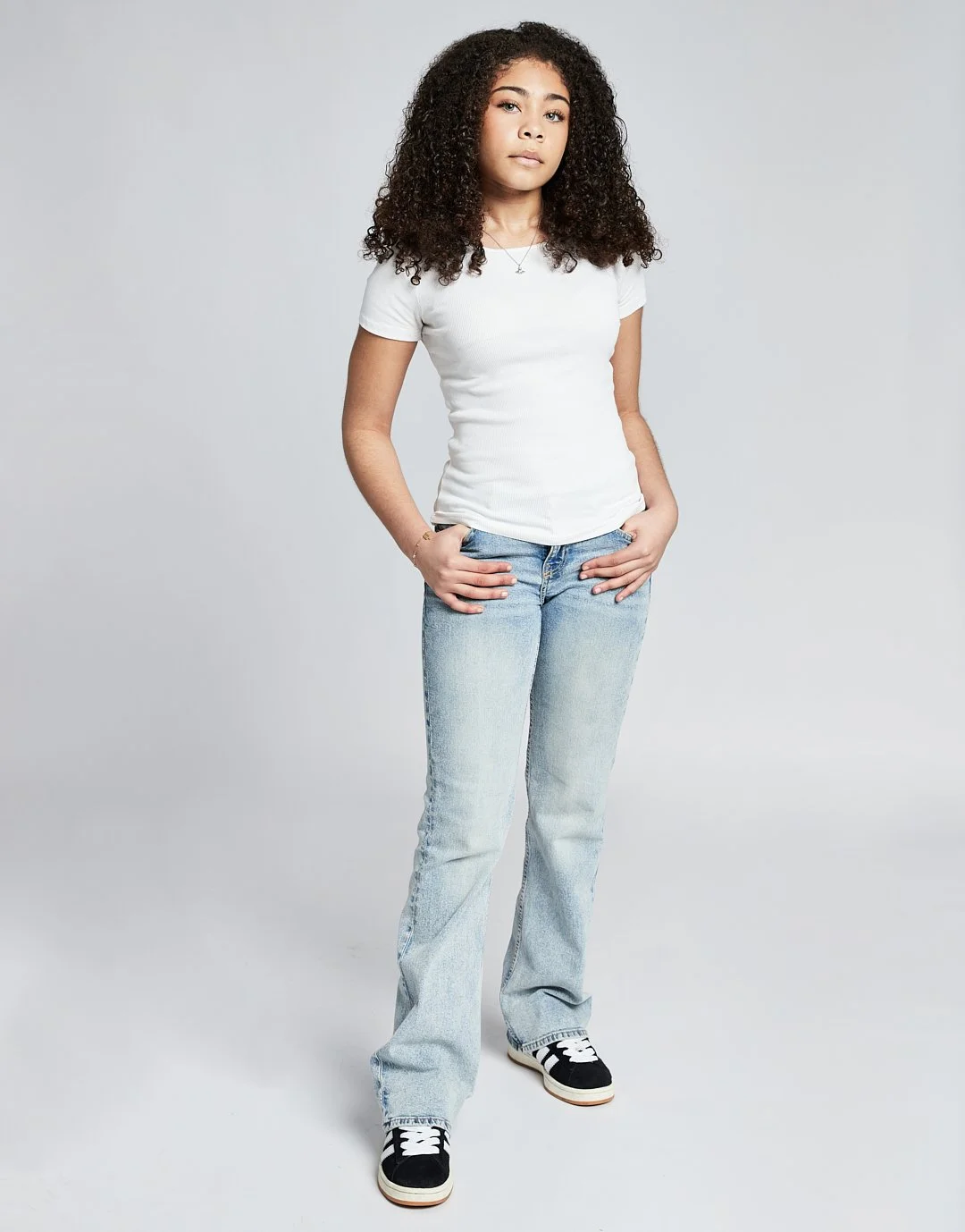 Young girl with curly hair wearing a white t-shirt, light blue jeans, and black and white sneakers, standing against a plain gray background.