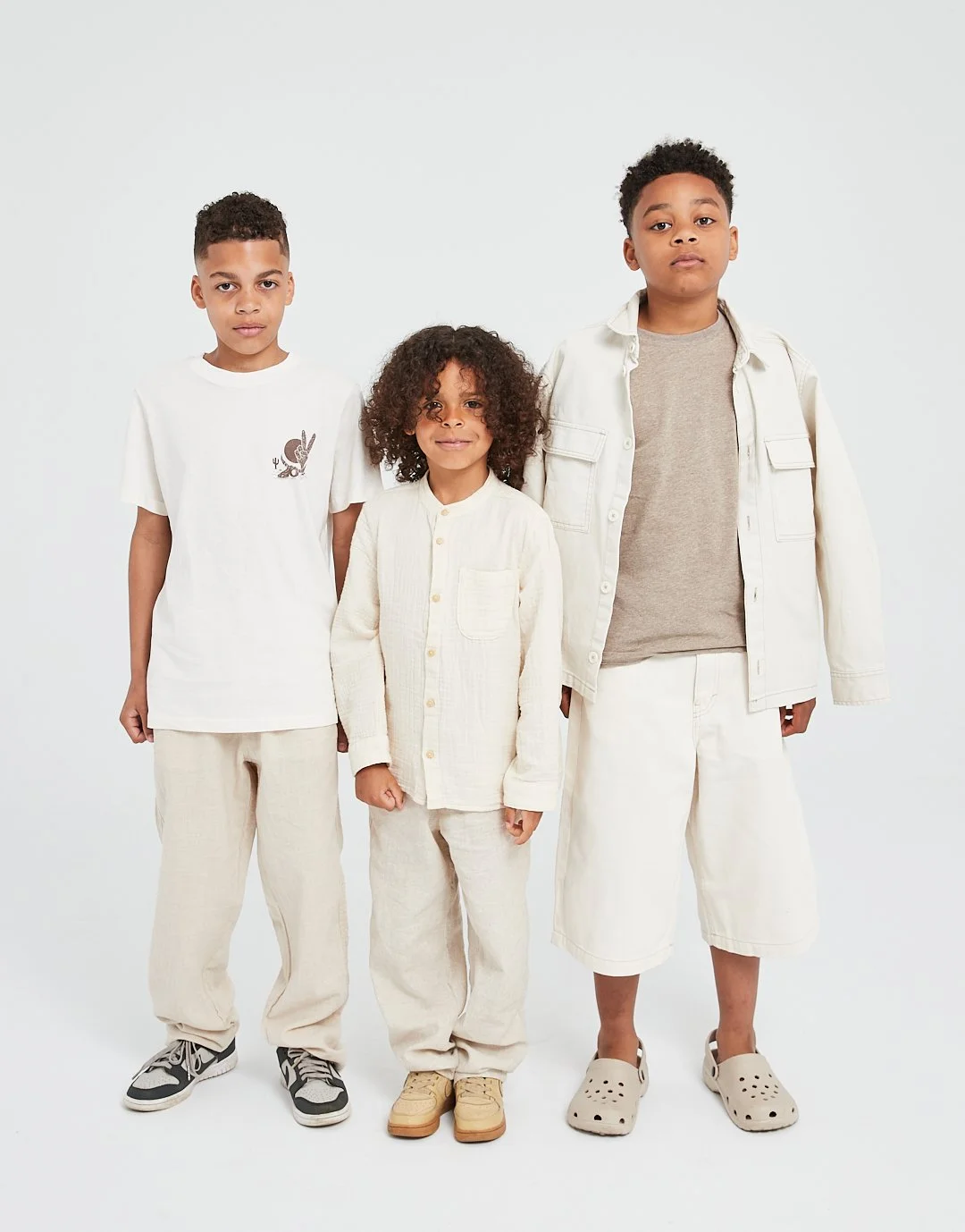 Three boys standing together in casual, neutral-colored clothing against a plain background.