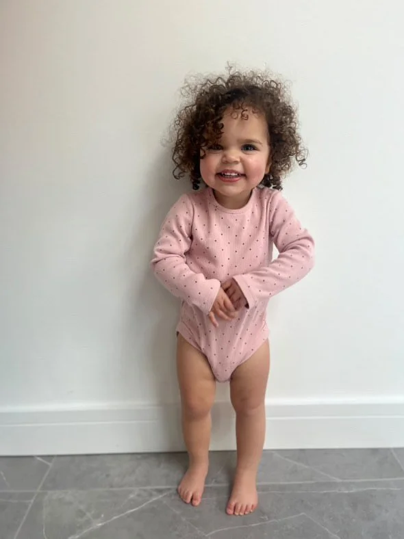 A young girl with curly hair wearing a pink long-sleeve shirt with tiny dots, standing barefoot against a white wall with a gray tile floor, smiling at the camera. Modelling for leading child model and talent agency SocityUK based in London and Manch
