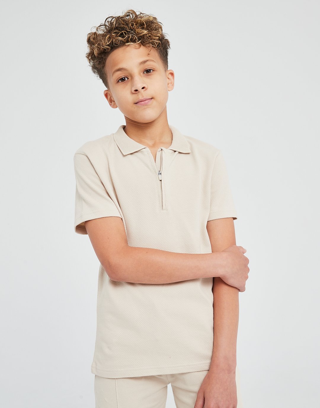 Young boy with curly hair wearing a beige polo shirt and matching pants, standing against a plain white background.