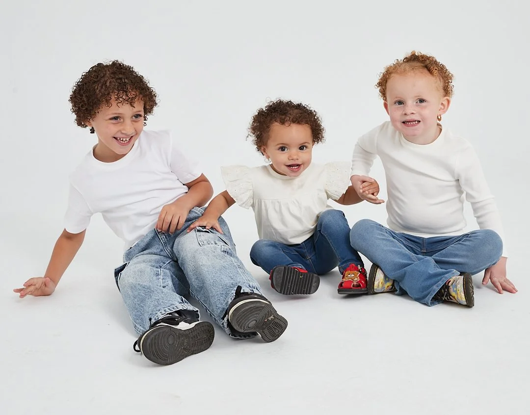 Three children sitting on the floor, smiling and holding hands, against a plain white background. Modelling for leading child model and talent agency SocityUK based in London and Manchester. Modelling for leading child model and talent agency SocityU