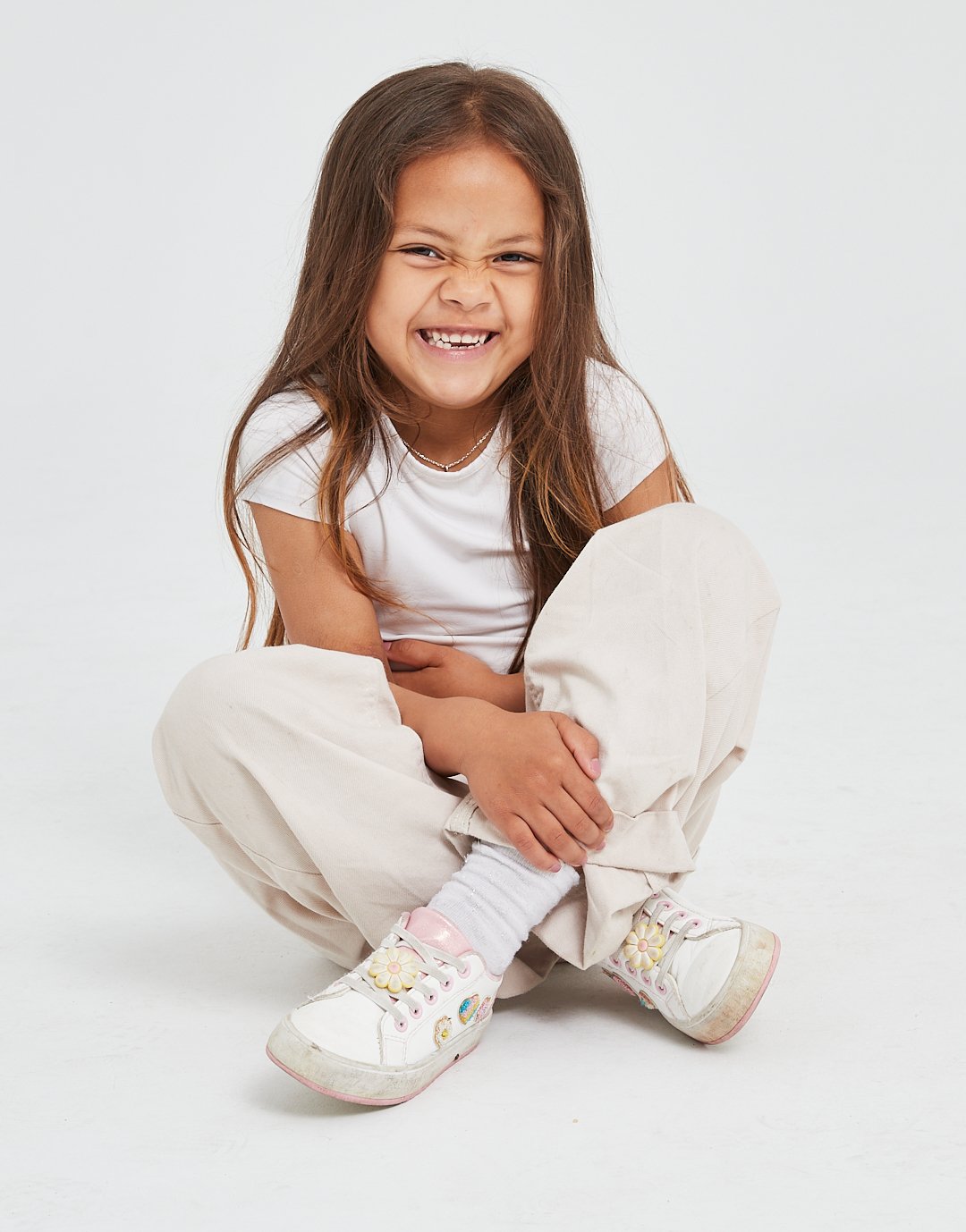 A young girl with long brown hair squatting, smiling with her teeth showing, holding her ankle, wearing a white t-shirt, beige pants, and white sneakers with colorful flower decorations.