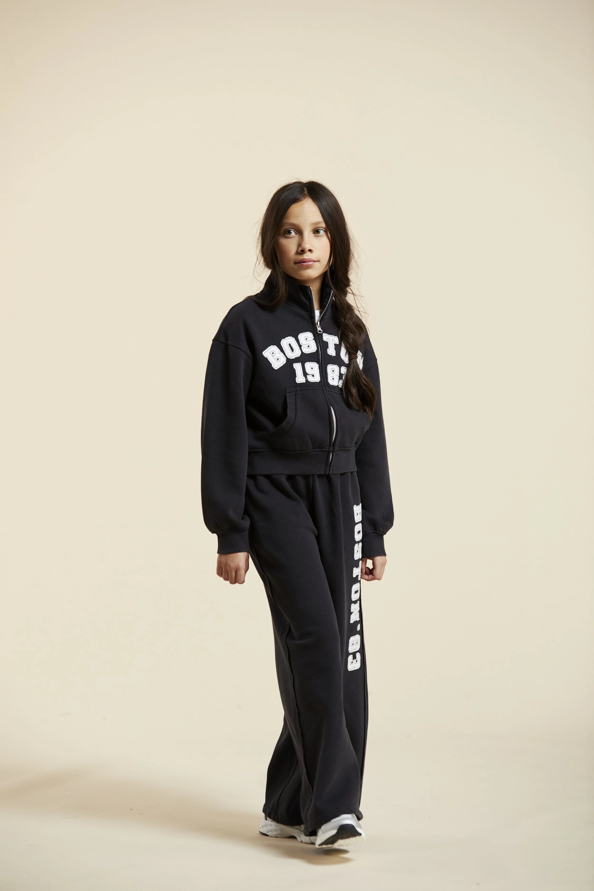 A young girl in a black athletic tracksuit with white lettering, standing against a plain beige background. Modelling for leading child model and talent agency SocityUK based in London and Manchester.