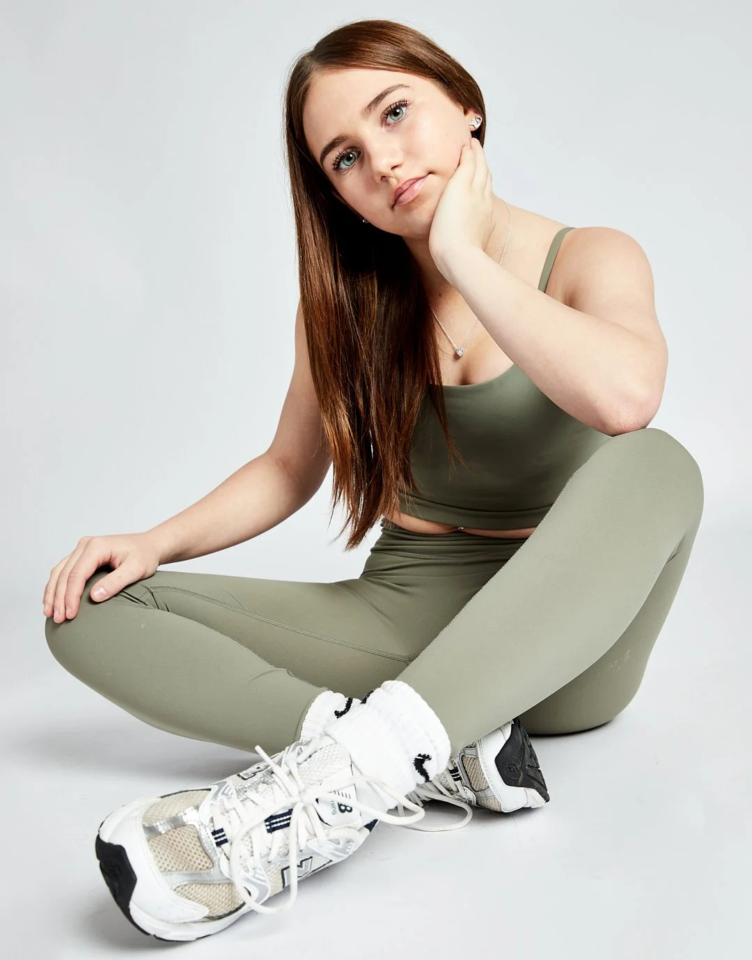 A young woman with long reddish-brown hair, wearing a matching olive green athletic crop top and leggings, white and beige sneakers, and white socks, sitting on the floor in a relaxed pose with her knees crossed. She rests her head on her left hand a