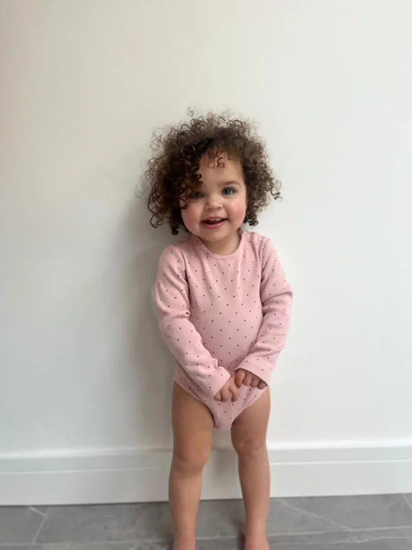 Little girl with curly hair wearing a pink polka dot long-sleeve shirt, standing against a white wall, smiling, with her hands clasped in front of her. Modelling for leading child model and talent agency SocityUK based in London and Manchester.