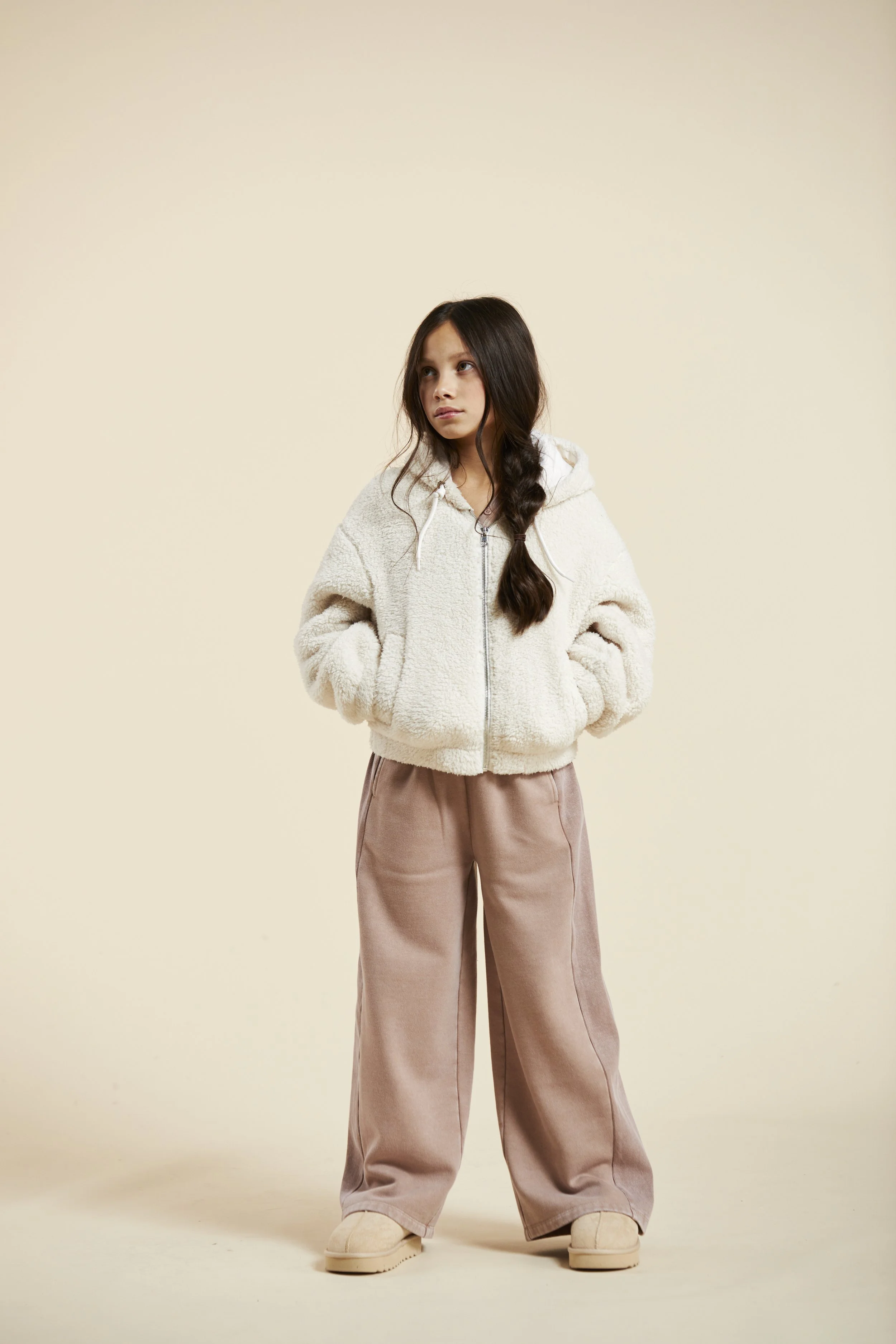 A young girl with long brown hair in a loose braid, wearing a fluffy cream-colored zip-up jacket with a hoodie and wide-bottom pinkish-brown pants, standing with her hands in her jacket pockets against a plain beige background. Modelling for leading 