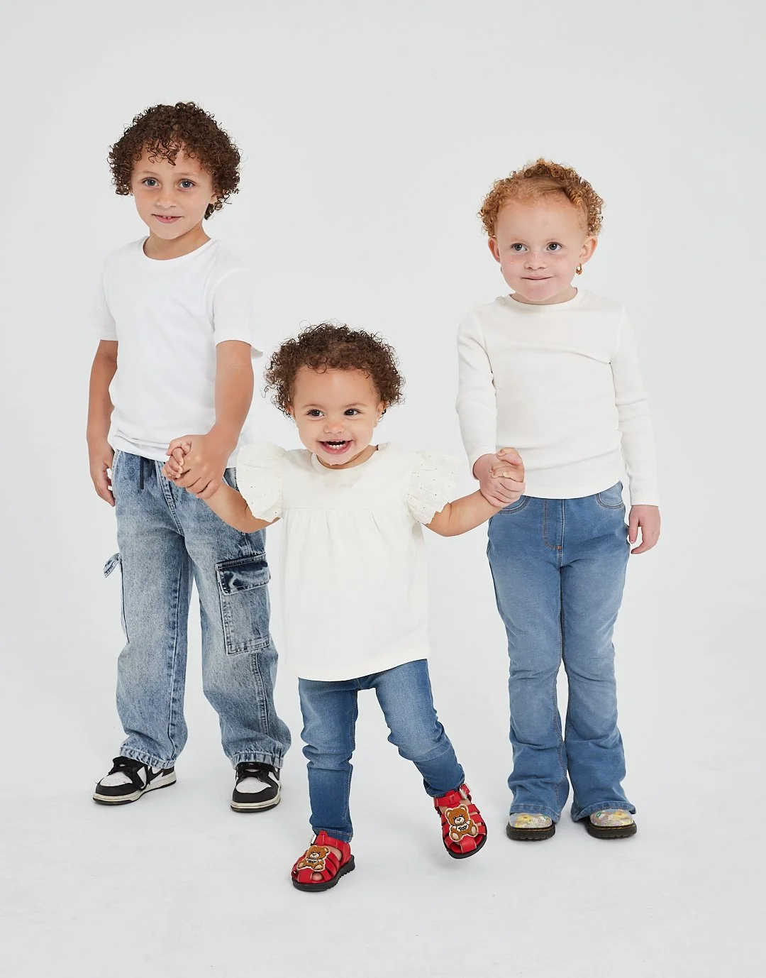 Three children holding hands, two standing on each side and one in the middle, all wearing white tops and blue jeans against a plain white background. Modelling for leading child model and talent agency SocityUK based in London and Manchester.