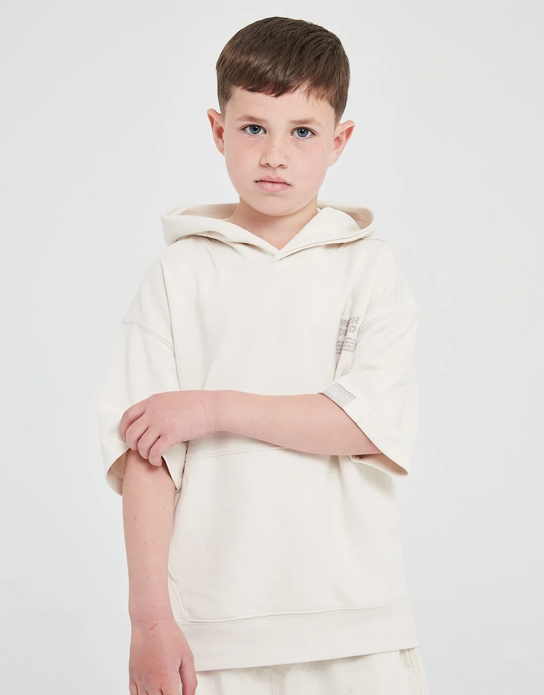 Young boy with short brown hair and blue eyes, wearing an oversized cream hoodie, standing against a plain white backdrop. He has a serious expression and his arms are crossed, with one arm slightly flexed to show a visible scar on his inner elbow.