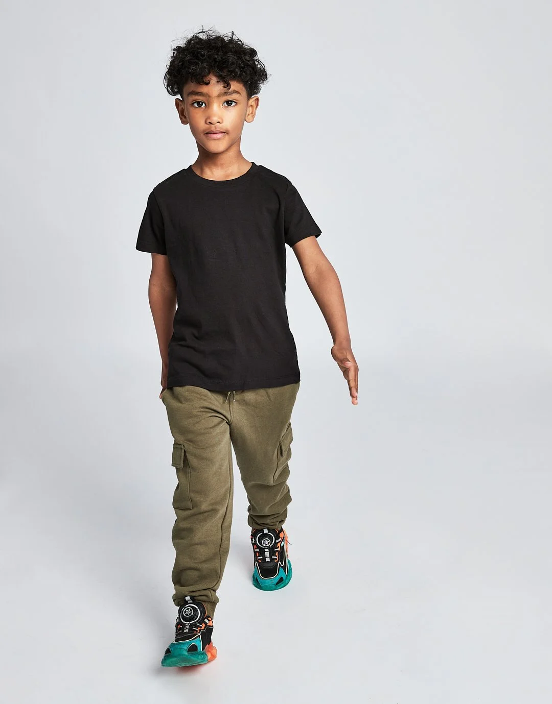 Young boy walking forward against a plain white background, wearing a black t-shirt, green cargo pants, and colorful sneakers.Modelling for leading child model and talent agency SocityUK based in London and Manchester.