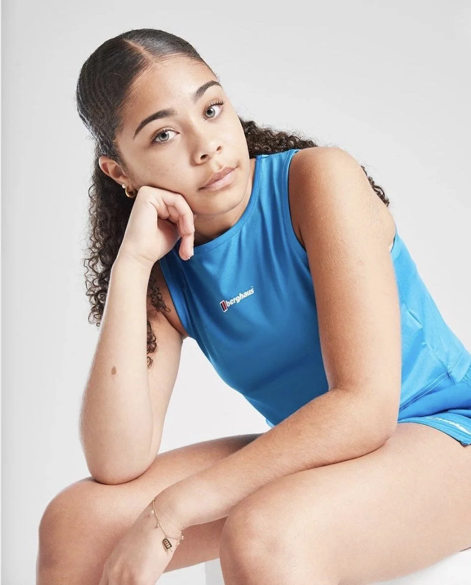A young woman with curly hair, wearing a blue sleeveless top, sitting with her chin resting on her hand, looking at the camera against a plain white background.
