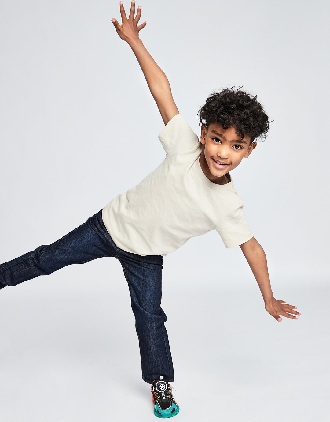A young boy with curly hair is balancing on one leg, leaning against the ground with his left hand, and raising his right arm in the air, against a plain white background. Modelling for leading child model and talent agency SocityUK based in London a