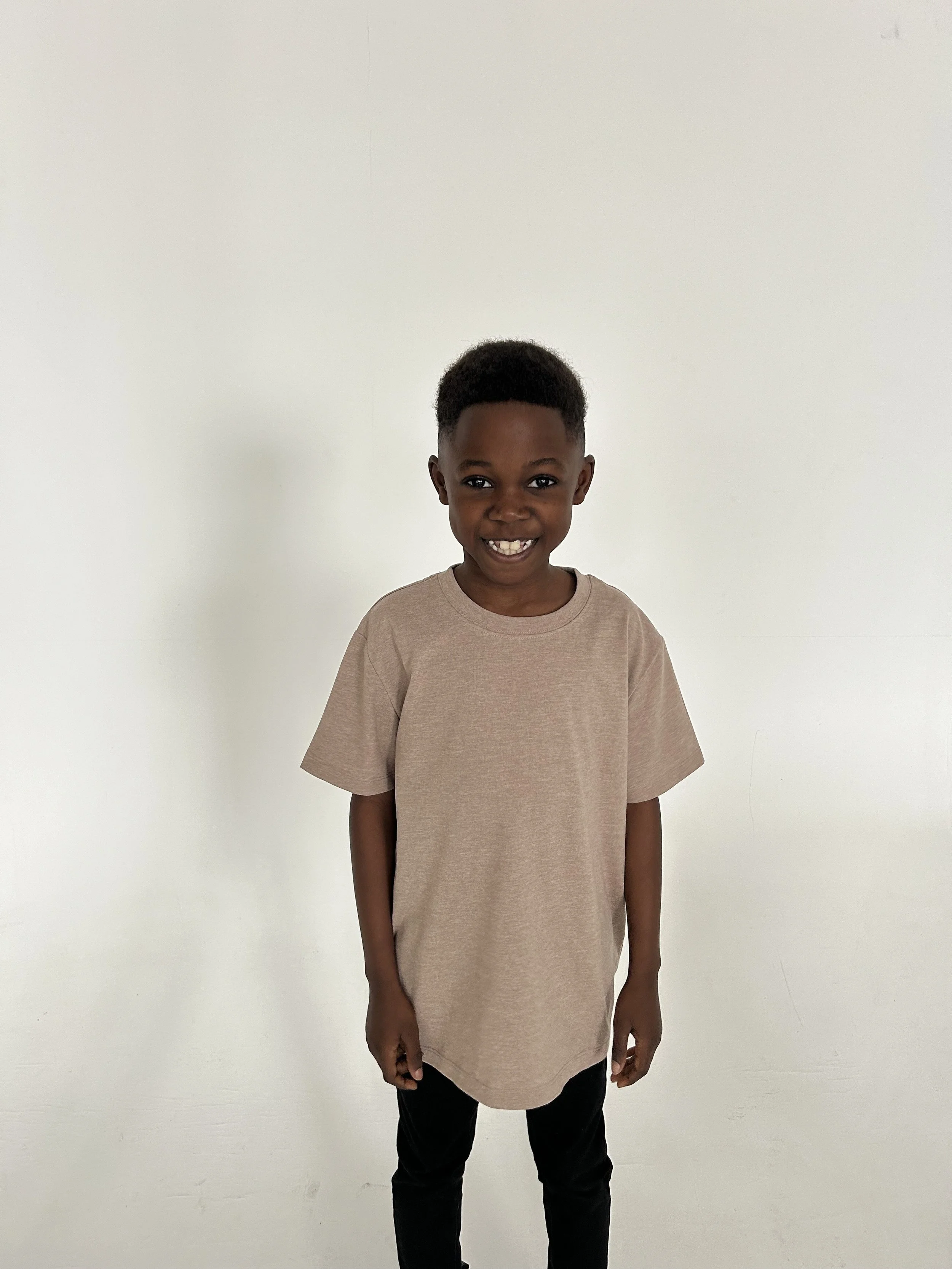 A young boy standing against a plain white wall, smiling and wearing a beige T-shirt and black pants. Model for leading Manchester and London based child model agency Socityuk.