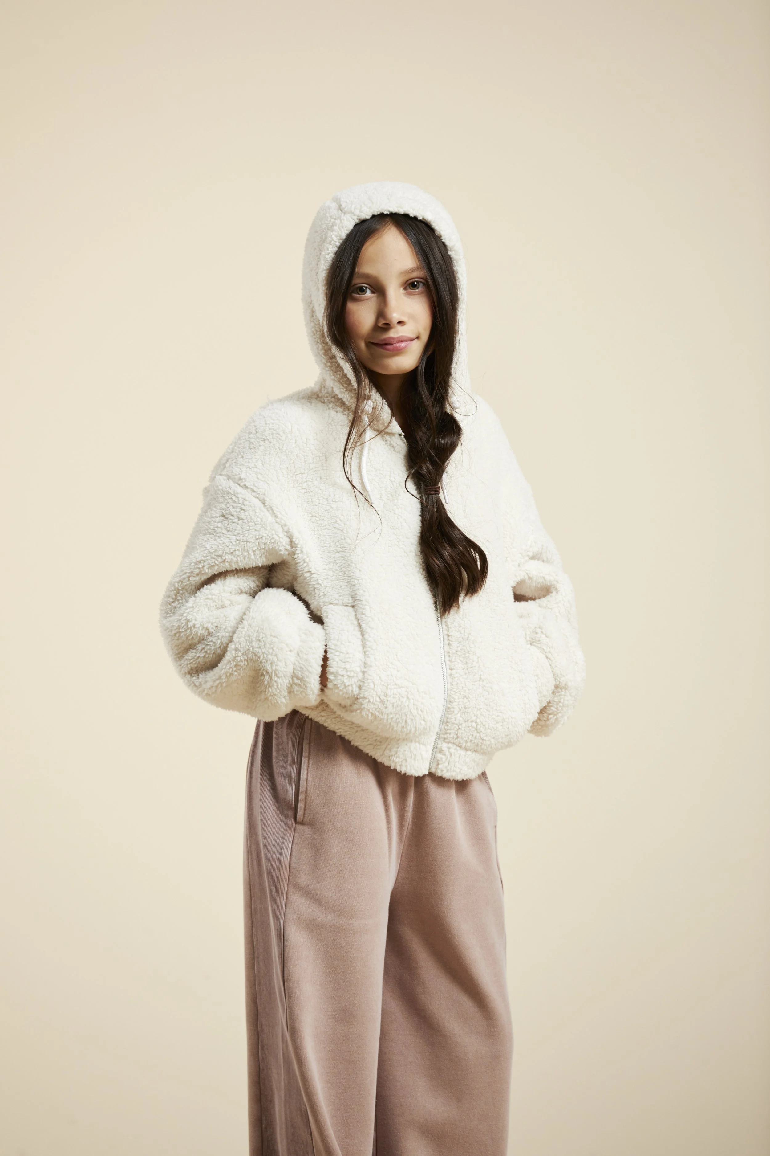 A young girl wearing a white fluffy hooded jacket and beige pants, standing with hands in pockets against a plain beige background. Modelling for leading child model and talent agency SocityUK based in London and Manchester.