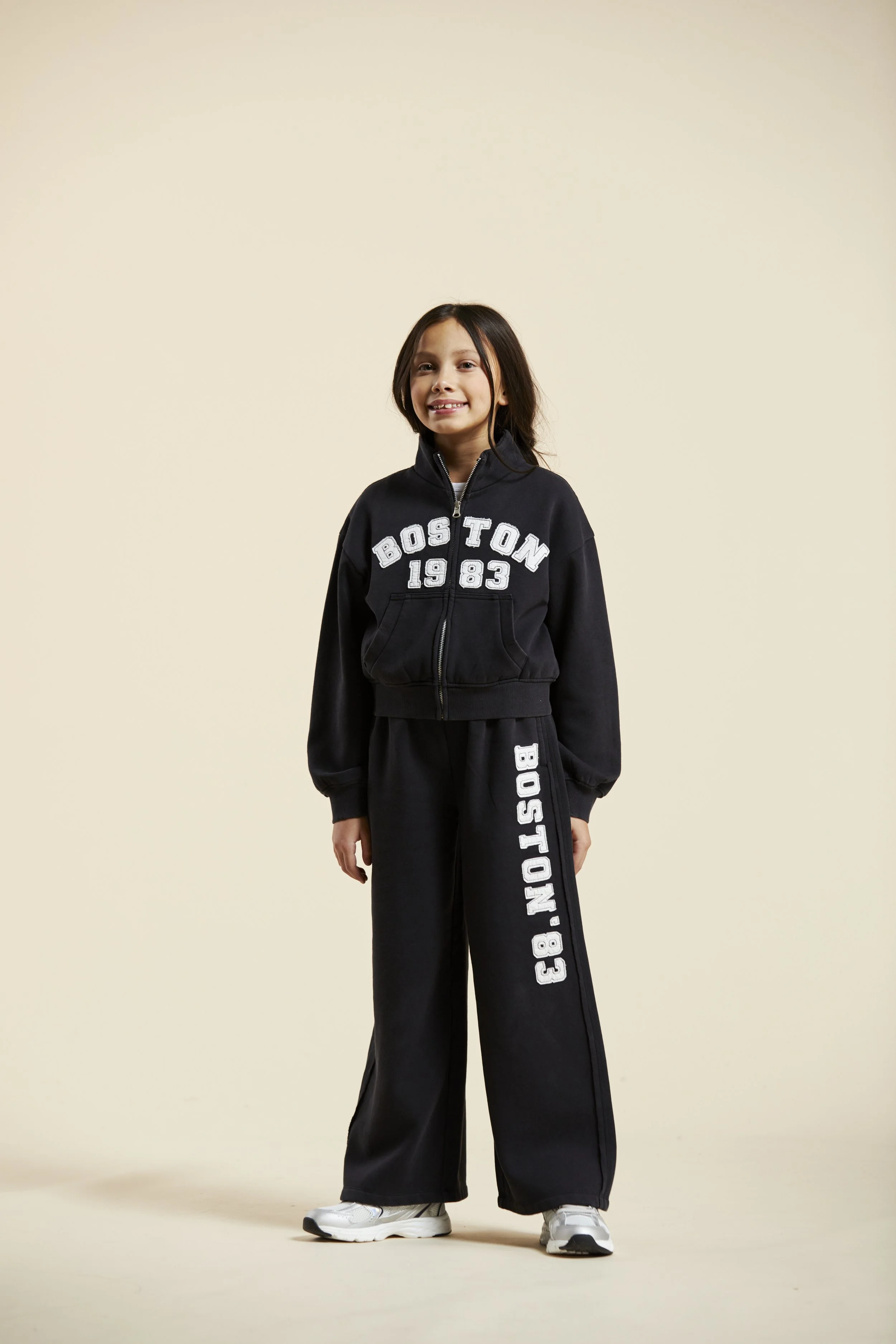 A young girl with long dark hair is dressed in a black sporty tracksuit with 'Boston 1983' printed in white on the jacket and pants, standing against a plain beige background. Modelling for leading child model and talent agency SocityUK based in Lond