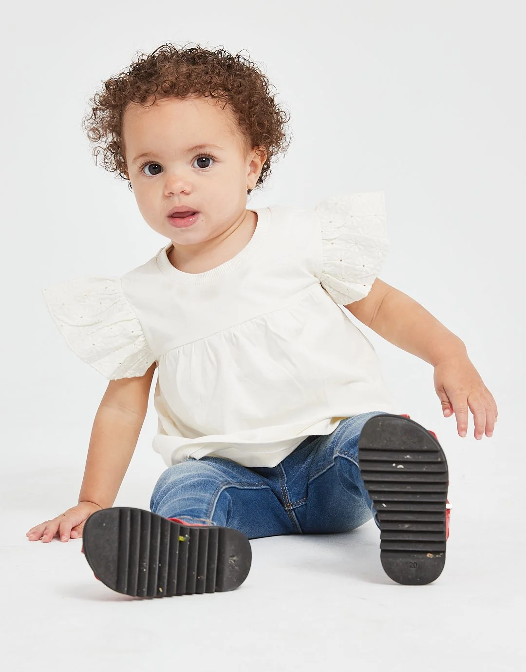 Young child with curly hair wearing a white top with puffed sleeves and blue jeans, sitting on the floor with legs spread and looking at the camera. Modelling for leading child model and talent agency SocityUK based in London and Manchester.