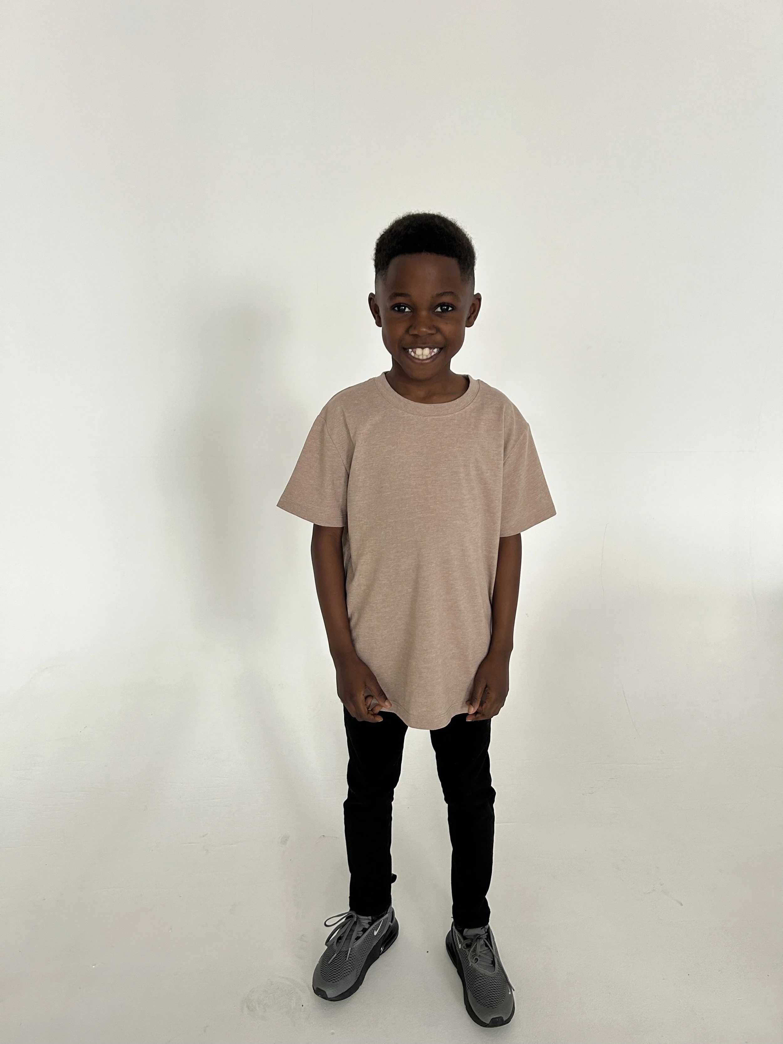 Smiling young boy with dark skin, short curly hair, wearing a beige oversized T-shirt, black pants, and gray sneakers standing against a plain white background. Model for leading Manchester and London based child model agency Socityuk.