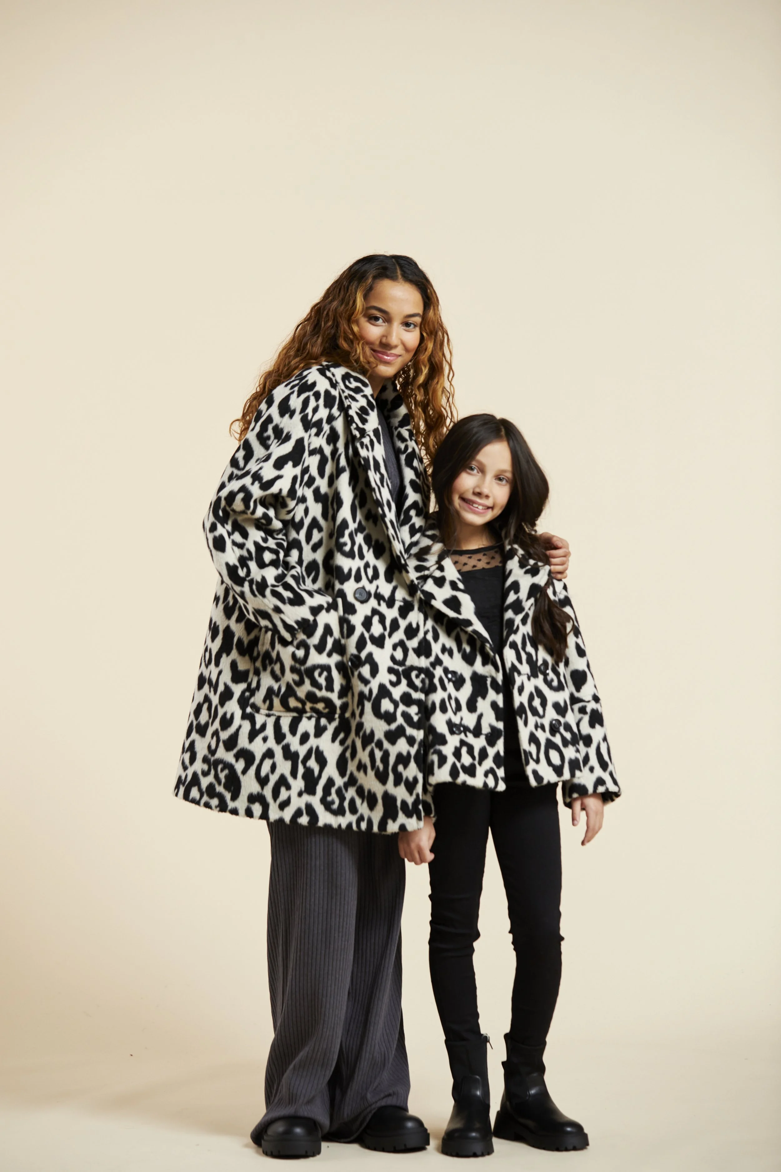 A woman and a girl standing close together, both wearing matching black and white animal print coats, smiling at the camera against a plain beige background. Modelling for leading child model and talent agency SocityUK based in London and Manchester.