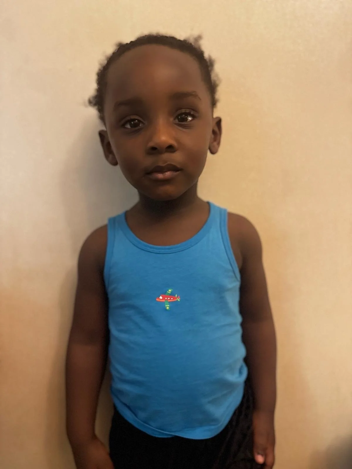 A young boy with dark skin and black hair styled in small twists, wearing a blue sleeveless shirt with a small colorful airplane graphic on the chest, standing against a plain beige wall. Modelling for leading child model and talent agency SocityUK b