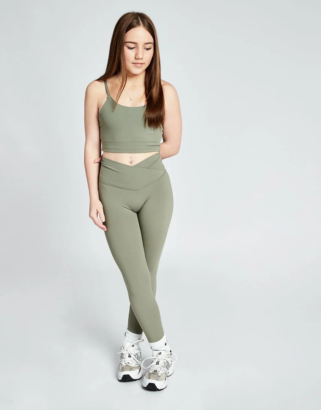 A young woman standing against a plain light gray background, dressed in olive green workout clothing, including a crop top and leggings, with white sneakers.